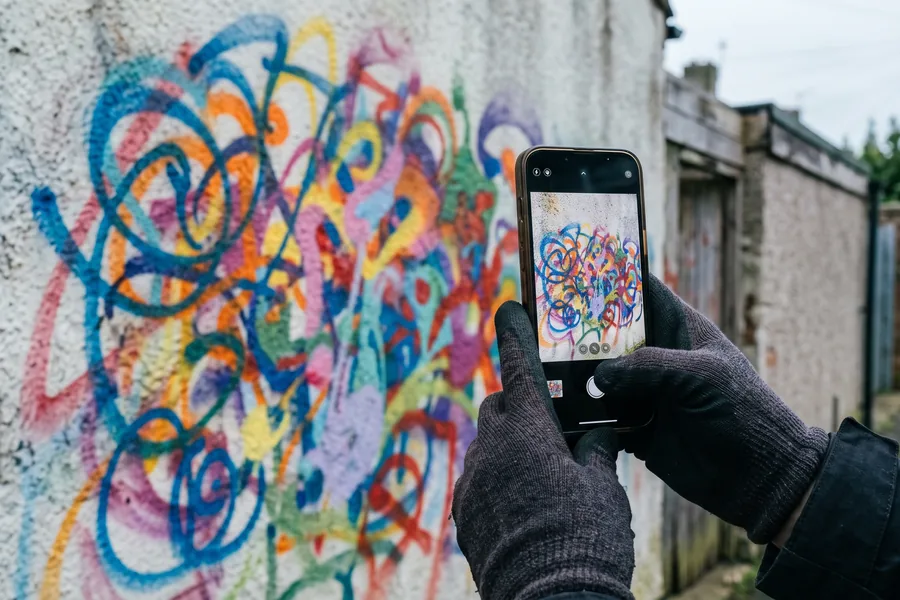 Smartphone in behandschuhter Hand fotografiert ein Graffiti an einer Hauswand zur Dokumentation